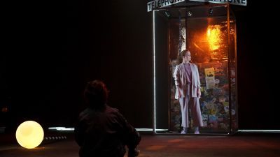 Actors on the sparse set of Residue under low lighting, one sits on the floor and the other stands in a telephone booth covered with stickers and posters on the inside